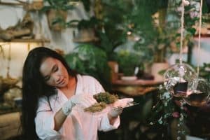 Woman in a white lab coat and gloves presenting a plate of mossy plant material in a lush greenhouse.