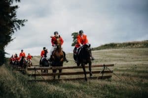 Group of riders in orange jackets jumping a wooden hurdle on an outdoor cross-country course.