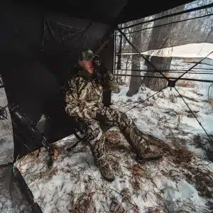 lQDPKcmUZ_kmSgPNBLDNBLCwjCpMbt8zxTMJJXuYzT_WAA_1200_1200 - North American Deer Hunter Man in camouflage sits inside a hunting blind in a snowy forest, looking through binoculars.