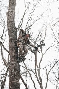 Hunter in camouflage climbs a tall tree in a snow-covered forest, holding a bow ready to shoot.