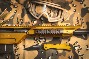 Close-up of a gold and black tactical rifle with a mounted rail, a yellow folding knife labeled 'Broken Skull', camouflage fabric background and protective goggles nearby.