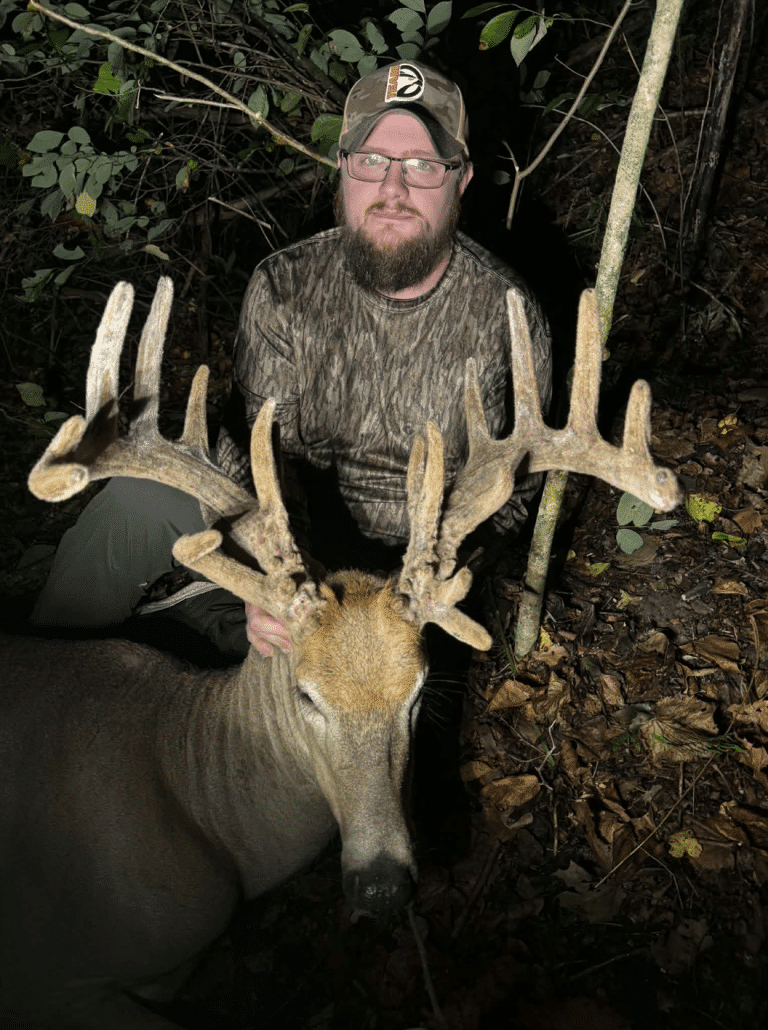 Hunter kneeling in camo at night in a forest, posing with a large buck and its tall, branched antlers.