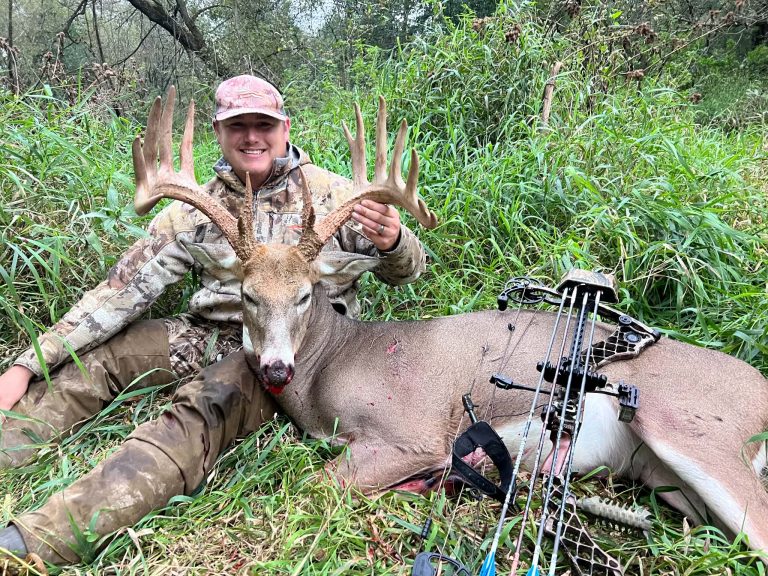 Camouflage-clad hunter holds up a large-antlered deer in tall grass with bow gear nearby.