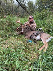 Hunter in camouflage sits among tall grasses posing with a large deer and its antlers, bow resting nearby.