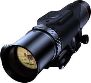 Black rifle scope viewed at an angle, with textured grip and adjustment knobs; the lens shows a deer standing in a grassy field.