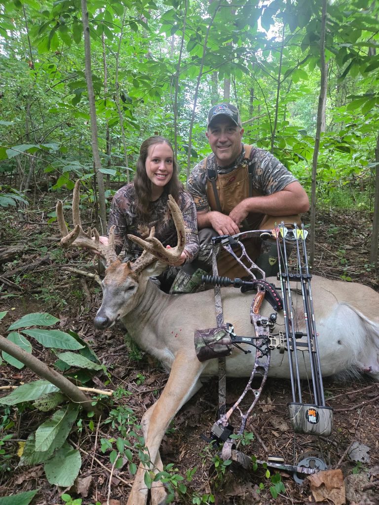 Two people in camouflage sit in a green forest with a large deer laid on the ground beside them, bow in the foreground.