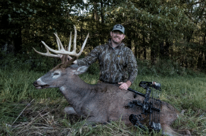 Hunter in camouflage posing with a large buck in a grassy forest clearing, bow beside him.