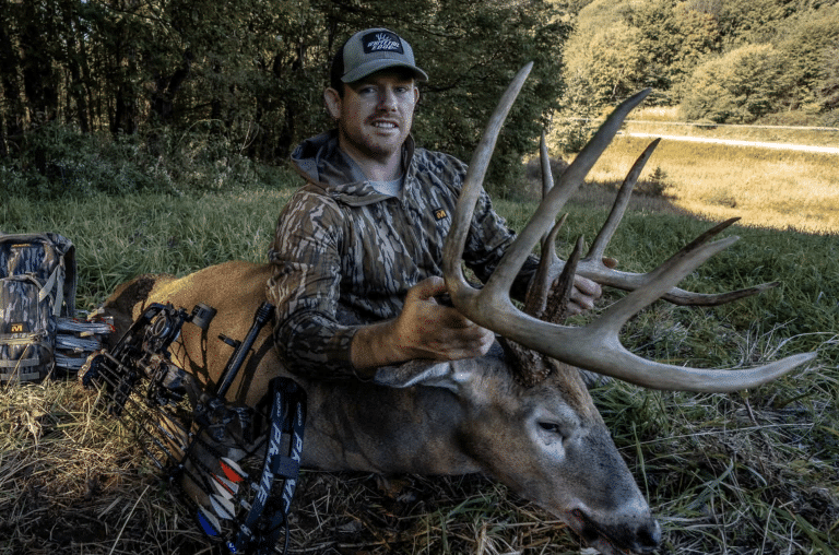 Hunter in camo jacket and cap holds a large deer by its antlers in a grassy field, bow and hunting gear nearby.