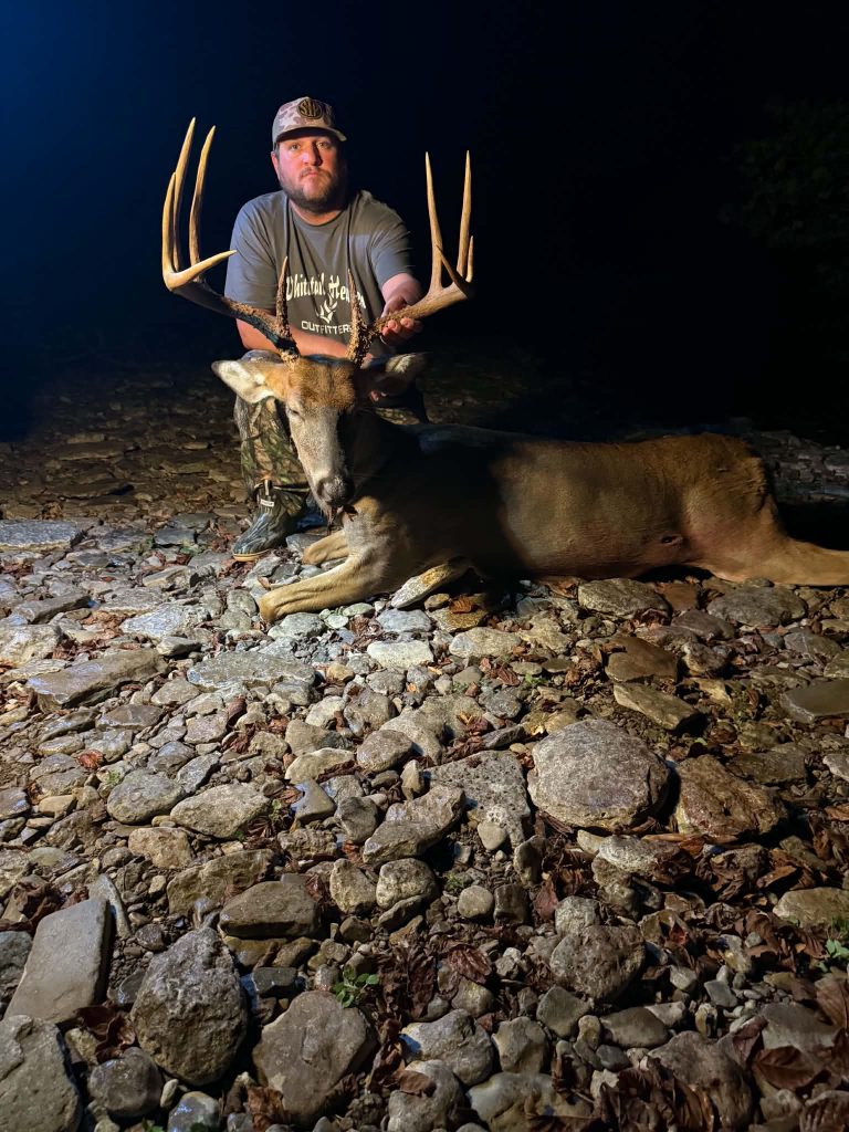 Man kneeling beside a large hunted deer with antlers on a rocky ground at night, illuminated by a strong light.