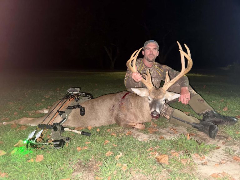 Hunter in camouflage at night kneels beside a harvested deer with large antlers, holding the antlers; bow and gear lie on the ground nearby.
